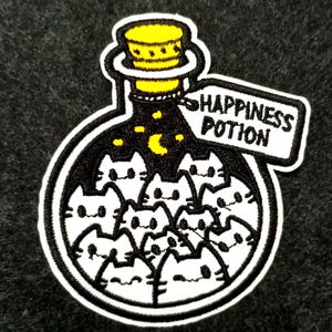May include: Embroidered patch featuring a black potion bottle filled with white cat faces. The bottle has a yellow cork and a tag that reads "HAPPINESS POTION". The background is black with yellow stars and a crescent moon.