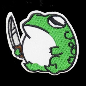 Può includere: Rana verde ricamata che tiene un coltello bianco. La rana è bianca con dettagli verdi e un occhio nero.