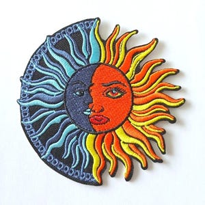 Puede incluir: Parche bordado con un diseño de sol y luna. El sol es naranja y amarillo con una cara humana, mientras que la luna es azul y negra con una cara humana. El parche está rodeado por un borde negro con un patrón de círculo repetido.