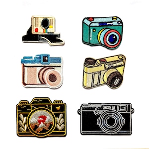 May include: Six embroidered patches featuring various camera designs. The collection includes a white instant camera, a teal camera, a blue and white camera, a yellow camera, a black camera with a mountain landscape lens, and a black camera.
