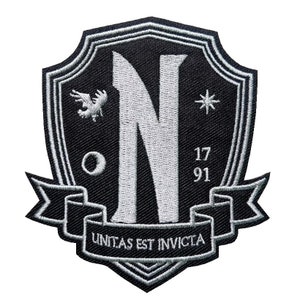 Può includere: Una toppa ricamata in bianco e nero a forma di scudo. Lo scudo presenta una grande lettera bianca "N", un piccolo uccello bianco, una stella bianca e i numeri "17 91". Un nastro bianco con il testo "UNITAS EST INVICTA" è avvolto attorno alla parte inferiore dello scudo.