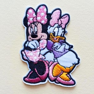 Puede incluir: Parche bordado con Minnie Mouse y Daisy Duck. Minnie Mouse lleva un lazo y un vestido de lunares rosas, mientras que Daisy Duck lleva un vestido y un lazo morados. Ambos personajes se abrazan, con un borde blanco.