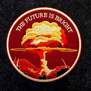 Op de afbeelding: Ronde geborduurde patch met een rode achtergrond en een witte rand. Het toont een gestileerde paddenstoelwolk in rood, oranje en geel, met de tekst "THE FUTURE IS BRIGHT" in het wit. Een uniek accessoire.