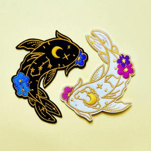 May include: Two embroidered patches shaped like koi fish. One patch is black with gold celestial patterns, a yellow crescent moon, and blue flowers. The other patch is white with gold celestial patterns, a yellow crescent moon, and purple flowers.