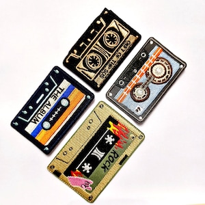 Op de afbeelding: Vier rechthoekige geborduurde patches in de vorm van vintage cassettebandjes. Eén is zwart met gouddraad, één is zwart met blauwe en oranje strepen en de tekst THE ALBUM, één is lichtblauw en grijs, en één is goudkleurig met de tekst ROCK.