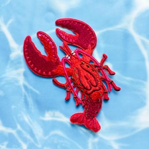May include: A red embroidered lobster patch with intricate detailing. The patch features a detailed design of a lobster with its claws and body. The background is a light blue with a water-like pattern.