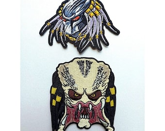 Op de afbeelding: Twee geborduurde patches met de Predator, een fictieve buitenaardse jager. De bovenste patch toont het hoofd van de Predator met dreadlocks, terwijl de onderste patch het gezicht van de Predator toont met een dreigende uitdrukking.