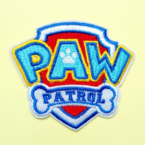 Może przedstawiać: Haftowana naszywka z logo Paw Patrol. Wzór zawiera napis PAW w kolorze jasnoniebieskim z białym odciskiem łapy oraz napis PATROL w kolorze niebieskim na białym tle w kształcie kości, a wszystko to na czerwonej tarczy herbowej.