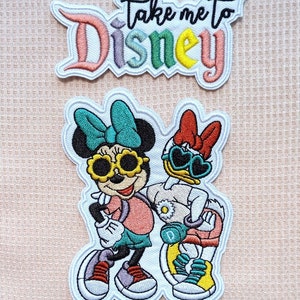 Puede incluir: Parche bordado para planchar con Mickey Mouse y Daisy Duck llevando gafas de sol y atuendos casuales. El texto "Take me to Disney" está en un parche separado.