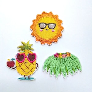 Op de afbeelding: Drie geborduurde strijkpatches met een gele zon met een zonnebril, een ananas met een zonnebril en een tropisch drankje, en een groene tropische bladerenrok met roze bloemen.