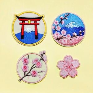 Könnte beinhalten: Vier bestickte Aufnäher mit japanischen Motiven: ein runder Aufnäher mit einem roten Torii-Tor und dem Berg Fuji, ein runder Aufnäher mit rosa Kirschblüten und dem Berg Fuji, ein runder Aufnäher mit einem Pflaumenblütenzweig und eine einzelne Blüte.