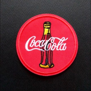 May include: A round, red embroidered patch featuring the Coca-Cola logo. The logo includes the Coca-Cola script in white and a stylized bottle with yellow and black details. The patch has a red border.