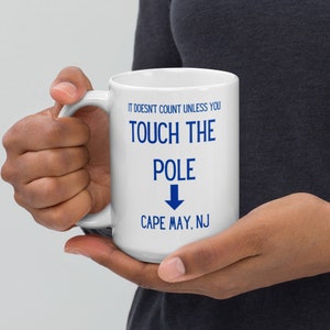 Puede incluir: Taza de café de cerámica blanca con texto azul que dice "It doesn't count unless you touch the pole Cape May, NJ" con una flecha azul apuntando hacia abajo.