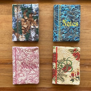 May include: Four small, quilted notebooks with embroidered "Notes" text. The notebooks feature various patterns, including floral, abstract, and a cat design. The colour palette includes green, blue, pink, and cream.