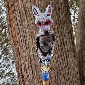 May include: A whimsical, hand-painted rabbit figurine with a distressed aesthetic. The rabbit has a gray head with red accents, a dark torso, and is mounted on a blue and gold staff. The staff features a crown emblem.