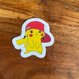 Puede incluir: Un personaje de dibujos animados Pikachu amarillo con una gorra de béisbol roja. La pegatina está sobre un fondo de madera.