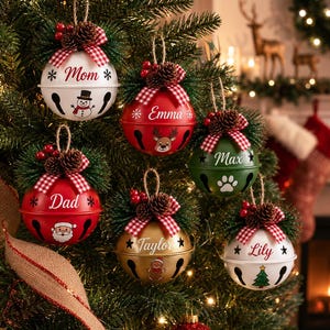 Six personalized Christmas jingle bell ornaments with custom family names and festive icons displayed in a gift box with pinecones, greenery and red gingham bows.