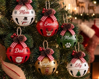 May include: Six round metal jingle bell ornaments hanging on a Christmas tree. Each ornament features a red and white checkered bow, pine needles, and small pinecones. They are personalized with names and festive graphics like snowmen, reindeer, Santa, paw prints, gingerbread men, and trees.
