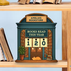 May include: A decorative bookstore-themed sign with the text "AMELIA'S BOOKSTORE" and "BOOKS READ THIS YEAR: 120." The sign features a green facade with a bookshelf interior, a potted plant, and a sleeping cat.
