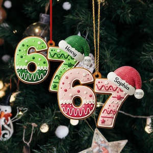 May include: Gingerbread-style Christmas ornaments shaped like the numbers 6, 7, and 7. The ornaments are decorated with white icing and feature names and the year 2025 on Santa hats. The ornaments hang from red and gold strings.