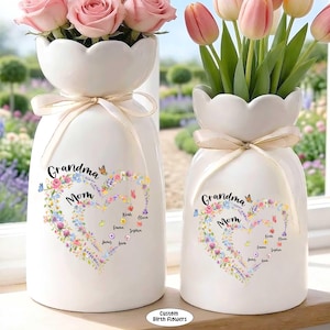 May include: Two white ceramic vases with scalloped tops, tied with beige ribbons, each holding flowers. One vase has pink roses, the other has pink and yellow tulips. Both vases feature a floral heart design with the words "Grandma Mom".