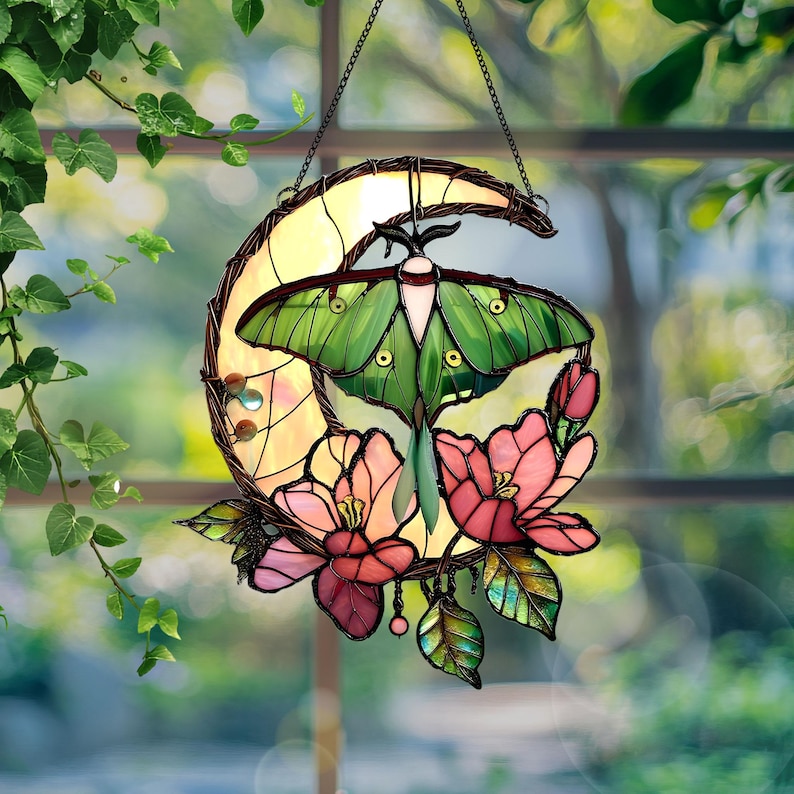May include: Stained glass suncatcher featuring a green moth with a cream-colored body, set against a yellow crescent moon. Pink and red flowers and green leaves adorn the bottom. The suncatcher hangs from a chain.