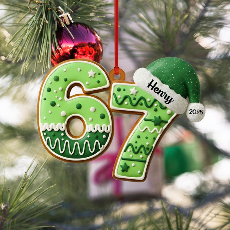 May include: A Christmas ornament shaped like the numbers 67, decorated with green icing and white accents. The number 7 is topped with a green Santa hat with the name "Henry" and the year "2025". The ornament hangs from a red ribbon.