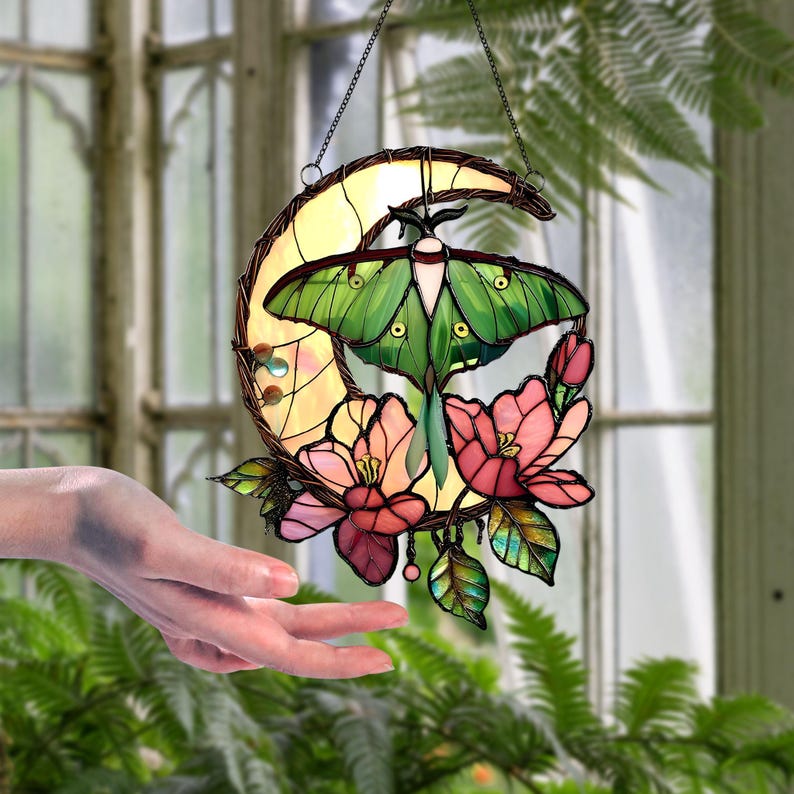 May include: Stained glass suncatcher featuring a green moth with yellow accents, set against a yellow crescent moon. Pink and red flowers and green leaves surround the moth and moon. The suncatcher hangs from a chain.
