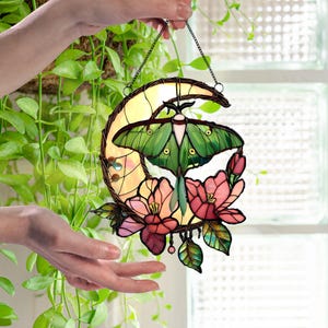 May include: Stained glass moon and moth suncatcher. The design features a green moth with pink and red flowers. The moon is yellow and the piece is framed with a dark metal. The suncatcher is hanging from a chain.