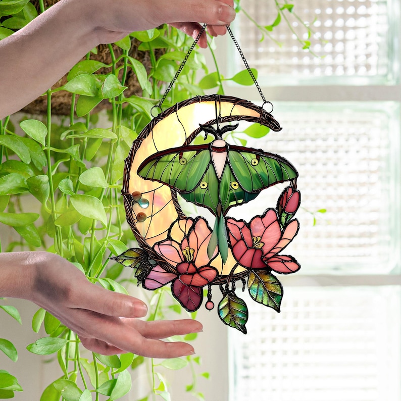 May include: Stained glass moon and moth suncatcher. The design features a green moth with pink and red flowers. The moon is yellow and the piece is framed with a dark metal. The suncatcher is hanging from a chain.