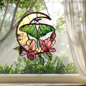 May include: Stained glass moon and moth suncatcher. The design features a green moth with pink flowers and leaves, set against a yellow crescent moon. The suncatcher is suspended by a chain and is designed to hang in a window.
