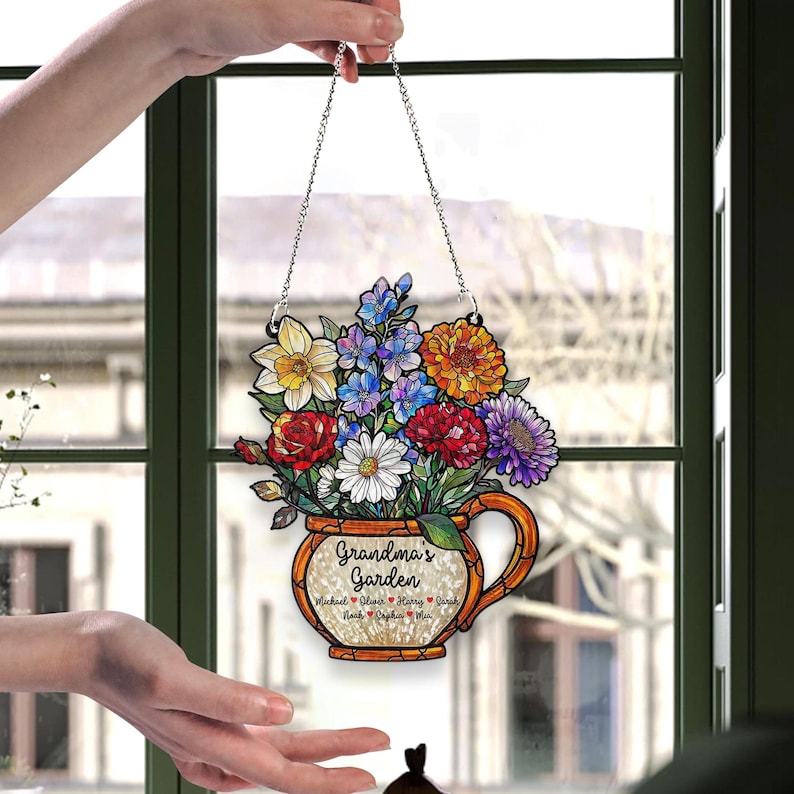 May include: A decorative hanging sign featuring a colorful floral arrangement in a decorative vase. The sign reads "Grandma's Garden" with names below. The flowers include yellow, red, purple, and white blooms. The sign is suspended by a silver chain.
