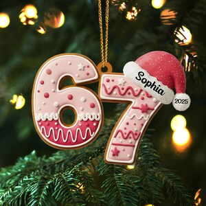May include: A festive Christmas ornament shaped like the number 67, decorated in pink icing with white and red accents. A small Santa hat with the name "Sophia" and the year "2025" is attached. The ornament hangs from a gold chain.