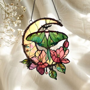 May include: Stained glass suncatcher featuring a green moth with detailed wings, set against a yellow crescent moon. Pink and red flowers and green leaves surround the moth. The suncatcher hangs from a chain.