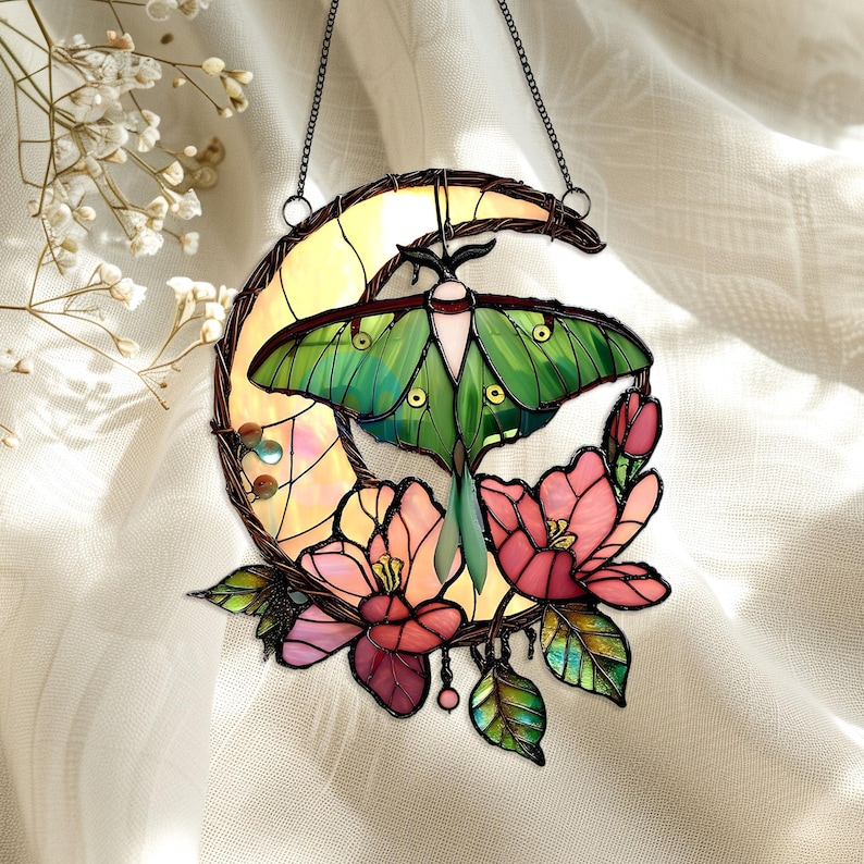 May include: Stained glass suncatcher featuring a green moth with detailed wings, set against a yellow crescent moon. Pink and red flowers and green leaves surround the moth. The suncatcher hangs from a chain.