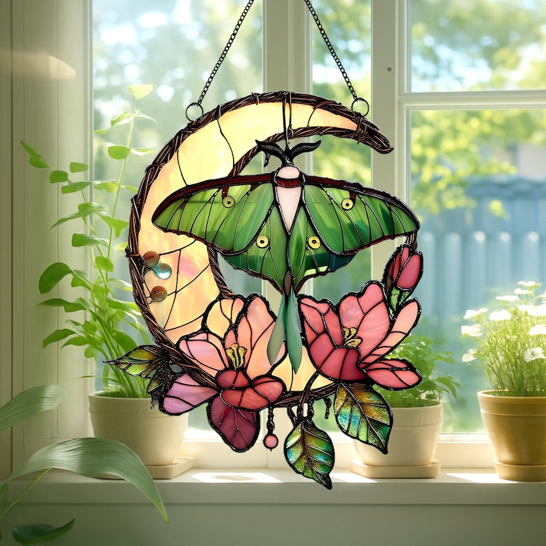 May include: Stained glass window hanging featuring a green moth with a cream-colored body and large green wings. The moth is set against a yellow crescent moon shape, with pink and red flowers and green leaves. The window hanging is suspended by a chain.