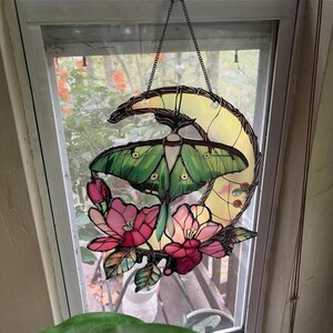 May include: Stained glass window hanging featuring a green moth with detailed wing patterns, set against a yellow crescent moon. Pink flowers and green leaves adorn the bottom. The piece is suspended by a chain.