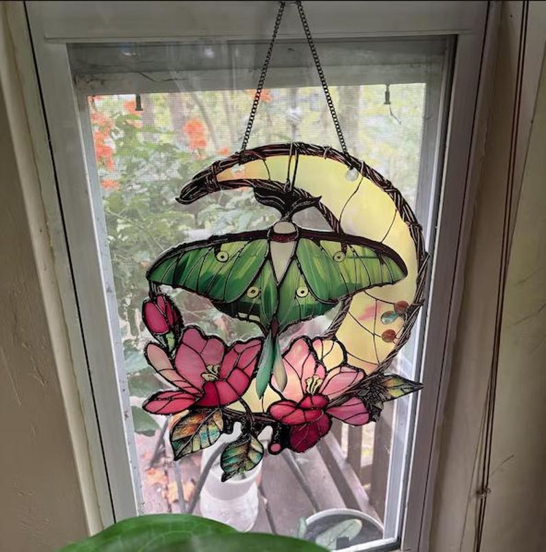 May include: Stained glass window hanging featuring a green moth with detailed wing patterns, set against a yellow crescent moon. Pink flowers and green leaves adorn the bottom. The piece is suspended by a chain.