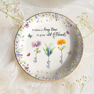 May include: A decorative white ceramic plate with a gold rim, featuring floral and butterfly designs. The plate has the text "It takes a long time to grow old Friends" and names Bella, Kaitlyn, and Charlie.