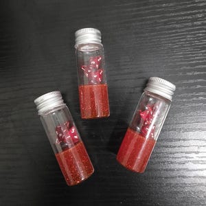 May include: Three small glass vials with silver screw-on lids. Each vial contains a set of red dice and a red glitter liquid. The vials are arranged on a dark wooden surface. The dice are visible through the clear glass.