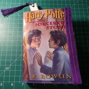 May include: A hardcover copy of Harry Potter and the Sorcerer's Stone by J.K. Rowling with a purple elastic band closure and a matching purple tassel bookmark. The cover features an illustration of a boy looking into a mirror.