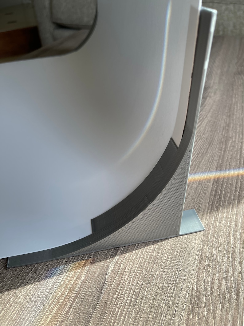 May include: A close-up of a gray plastic corner brace with a curved edge. The brace is attached to a white surface and is resting on a wood floor.