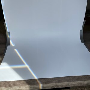 May include: A white backdrop for photography, set up on a wooden table. The backdrop is curved to create a seamless background. Sunlight is shining through a window, creating a rainbow effect on the backdrop.