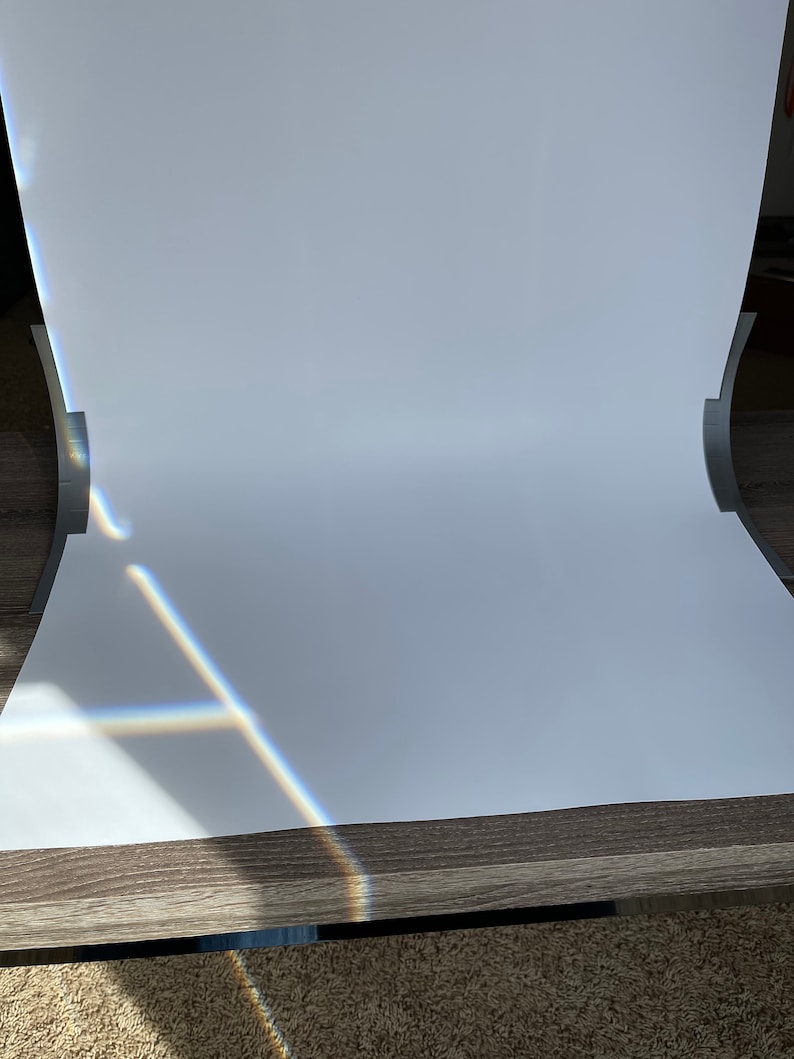 May include: A white backdrop for photography, set up on a wooden table. The backdrop is curved to create a seamless background. Sunlight is shining through a window, creating a rainbow effect on the backdrop.