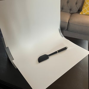 May include: A white backdrop with a black spatula and a black pen on a dark brown surface.