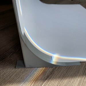 May include: A gray, curved backdrop stand with a white backdrop. The stand has a base with a rectangular shape and a curved support for the backdrop. The backdrop is slightly curved and has a slight sheen.