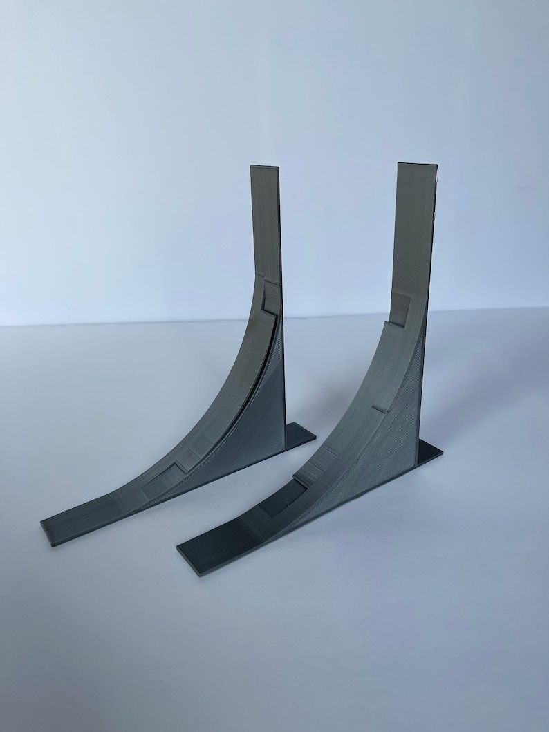 May include: Two gray 3D printed ramps, each with a curved surface and a flat base. The ramps are angled upwards and are positioned side by side.