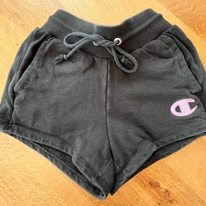 May include: Black athletic shorts with a pink Champion logo on the right leg. The shorts have a drawstring waistband and side pockets.