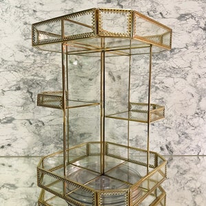 May include: A gold-toned, three-tiered, rotating glass organizer with a hexagonal base. Each tier has a glass shelf and a decorative beaded trim.