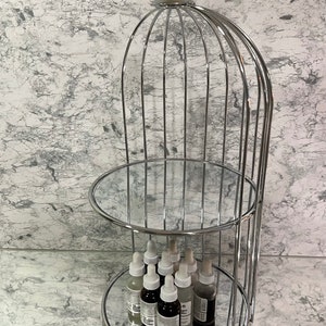 May include: A three-tiered, chrome-finished display stand with mirrored shelves. The top is adorned with a clear, crystal-like sphere. The stand holds several small, clear bottles with white droppers, likely skincare products. The background is a marbled wall.